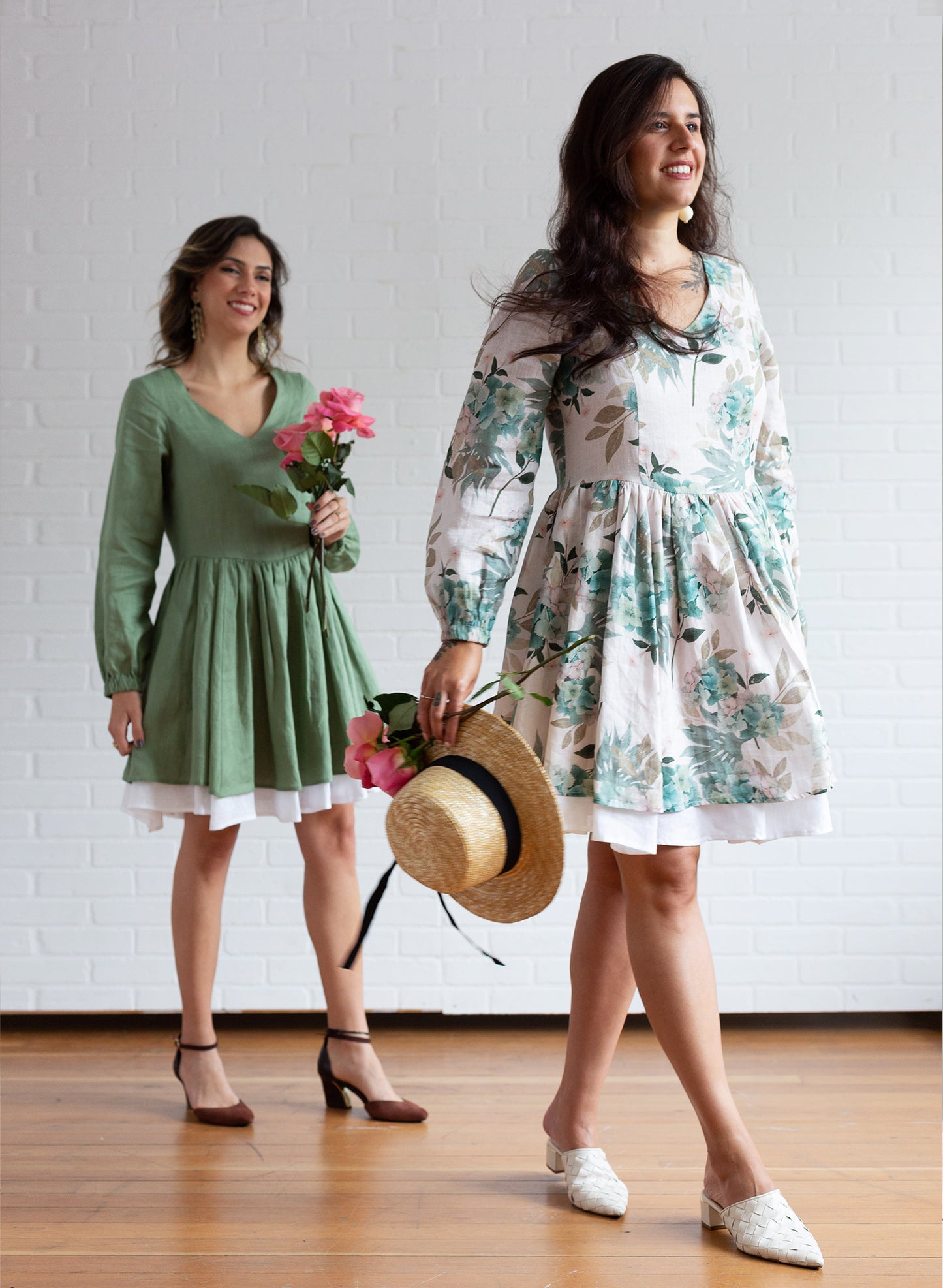 Two women wearing a green linen dress and a floral print linen dress holding flowers and a straw hat.